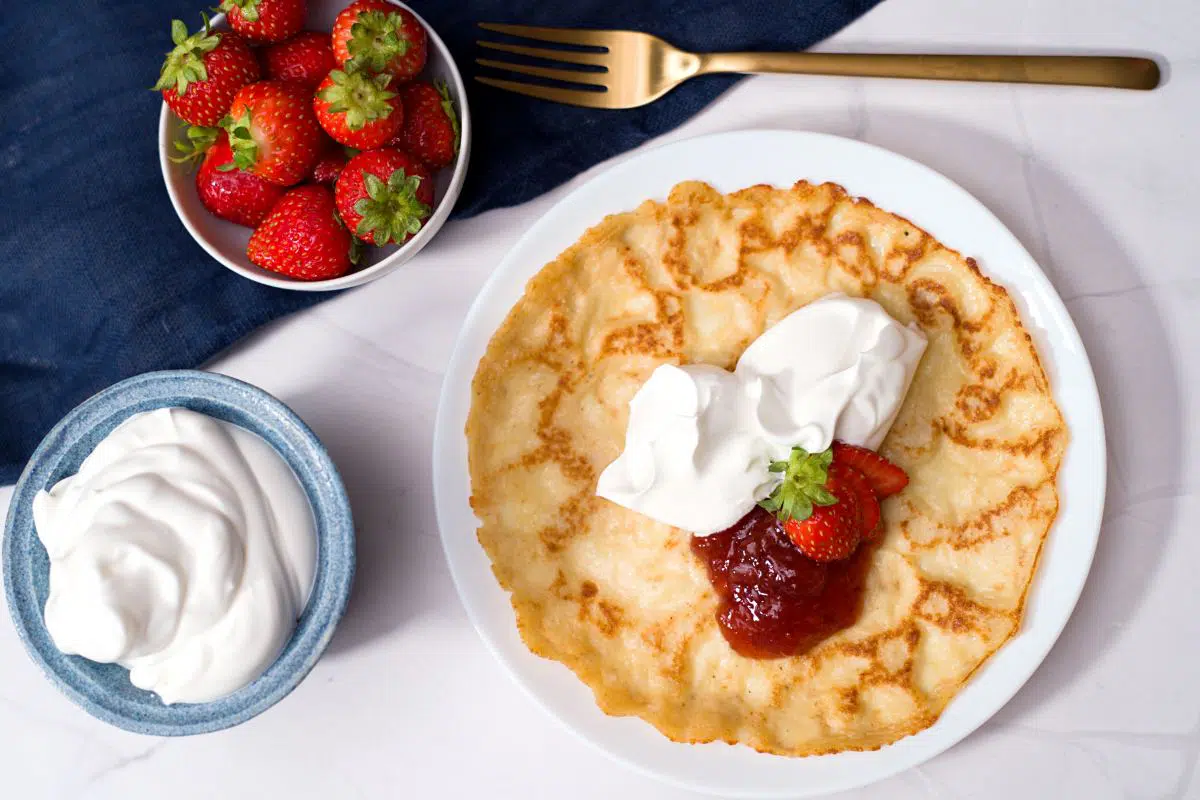 thin pancake served with whipped cream, strawberries and strawberry jam. In the background a blue napkin and the bowls for whipped cream and jam.