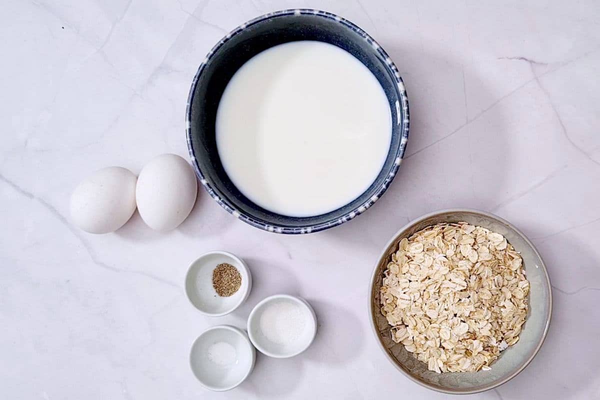 Milk, rolled oats, cardamom, sugar and salt in bowls and two eggs on table. 