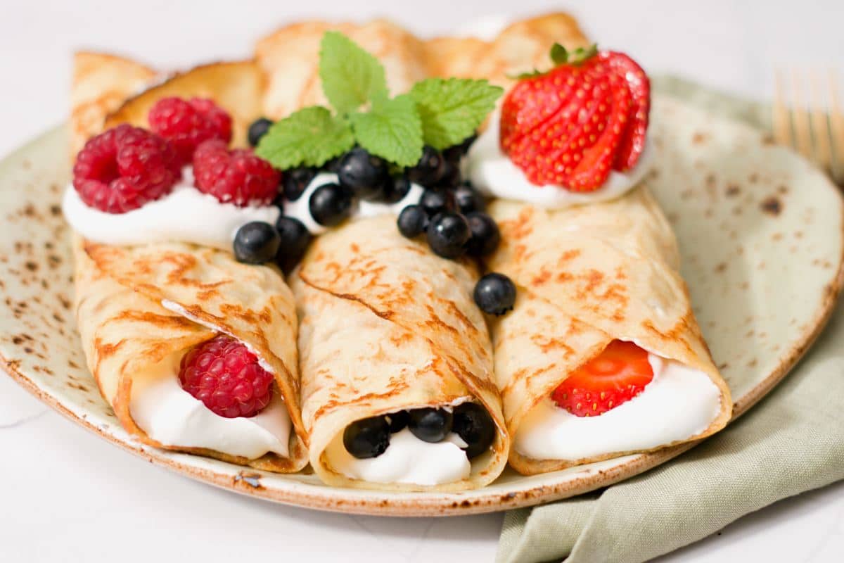 Three rolled pancakes on plate filled with whippped cream and decorated with fresh strawberries, blueberries and raspberries. 