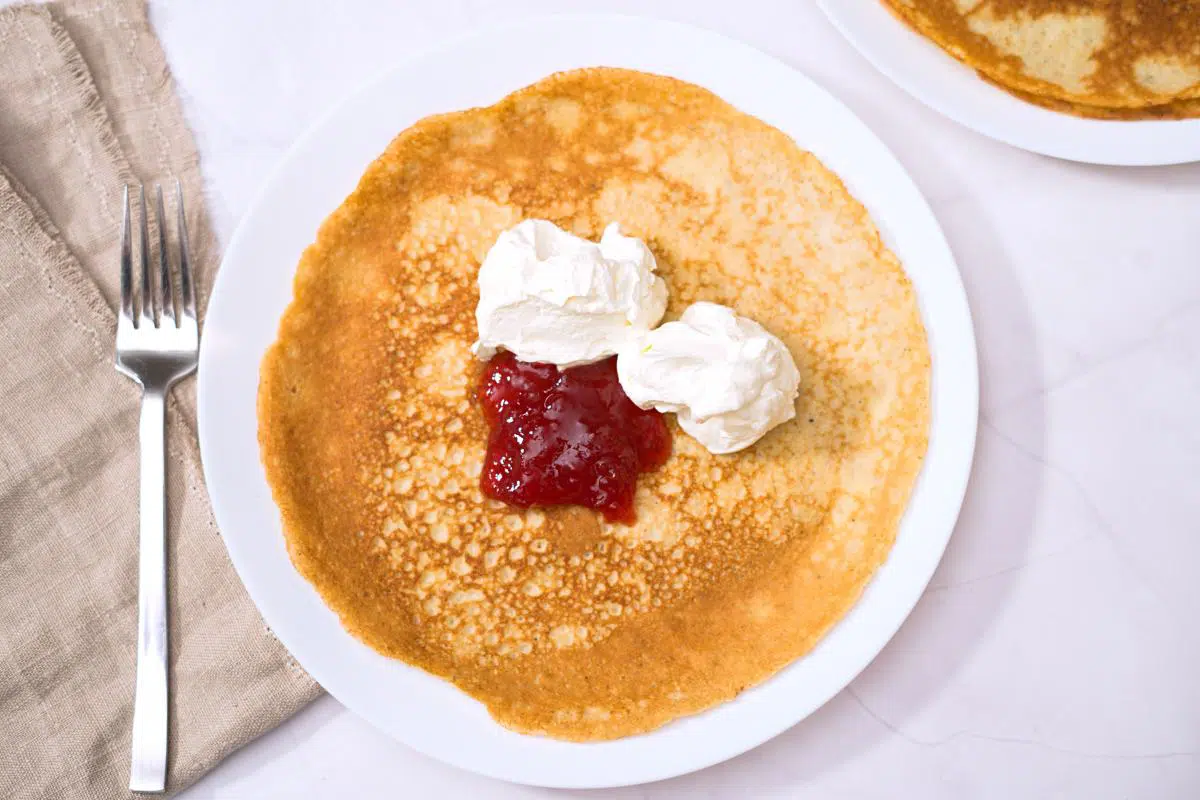 A thin pancake on white plate with strawberry jam and whipped cream. 