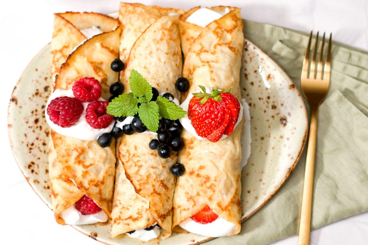 Three rolled pancakes on plate with filling and decorated with fresh berries, a golden fork and some mint leaves. 