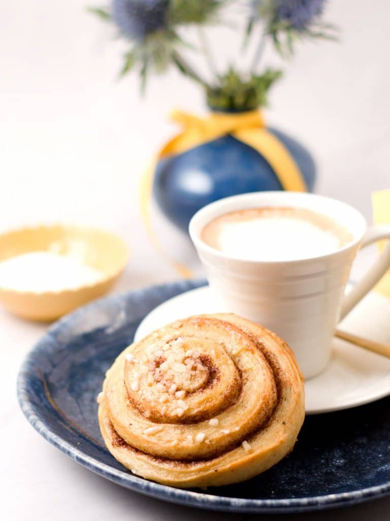 closeup of one cinnamon roll with a coffe cup and some flours and pearl sugar. 