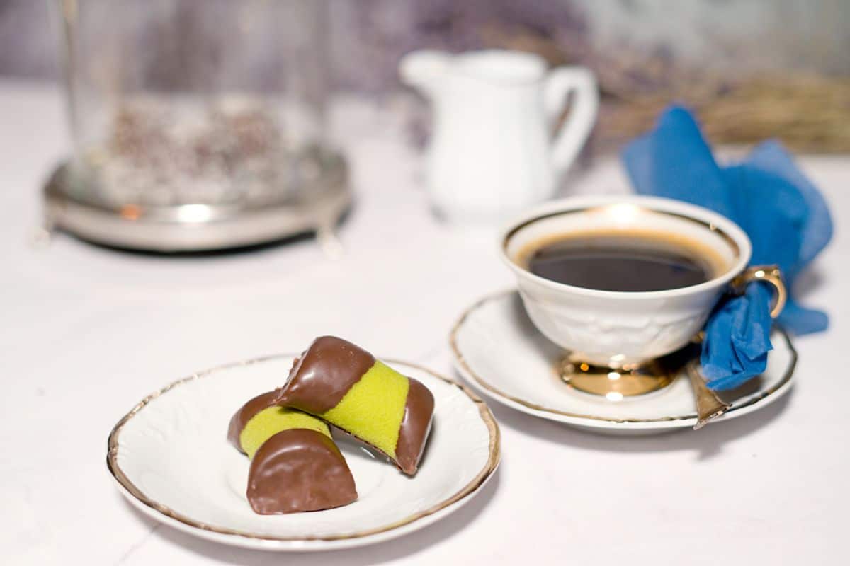A small coffee cup with napkin and two Swedish green dammsugare, marzipan-chocolate treats on a white small plate. 