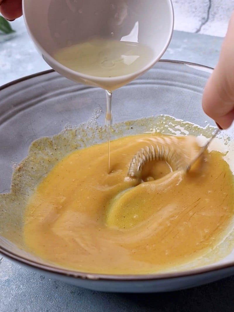 Pouring oil in bowl while whisking with a small hand whisk. 