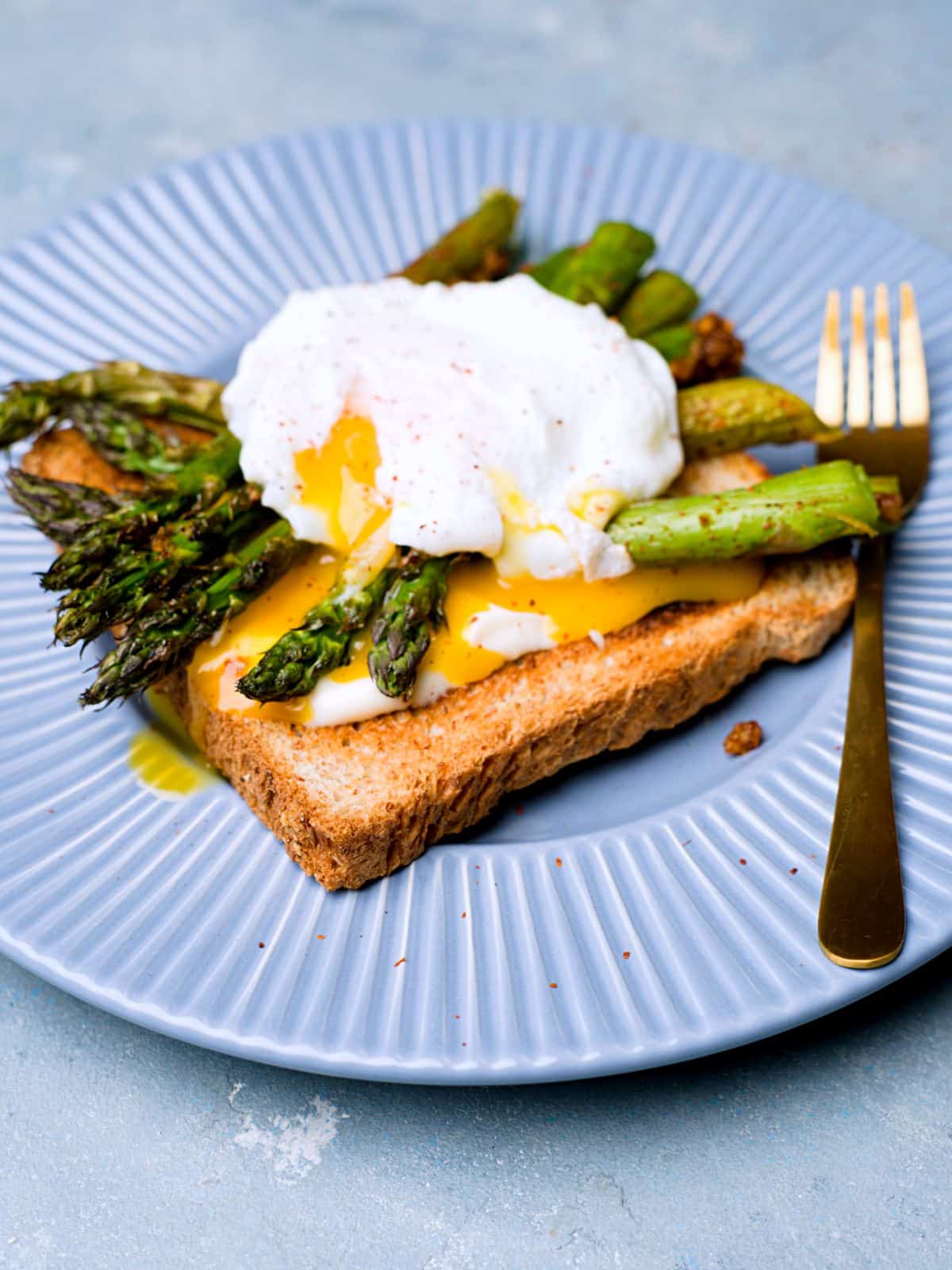 A toaste with roasted asparagus and a poached egg with egg yolk running out and a golden fork on blue plate, 