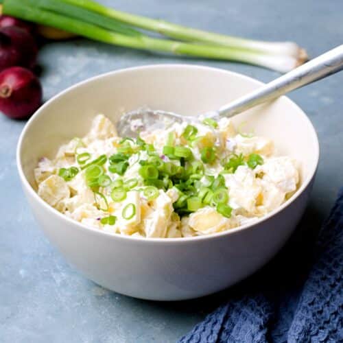 Large creamy potato salad in white bowl with red onions in background.