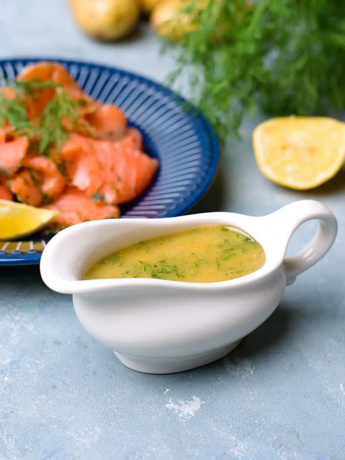 Yellowish mustard dill sauce in white pitcher with cured salmon plate in background. 