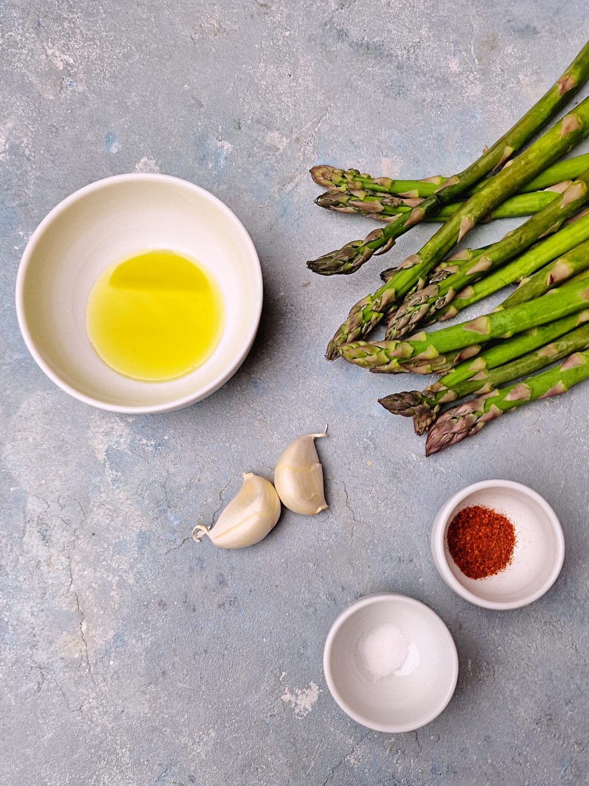 Asparagus, oil, garlic and spices in small bowls on surface. 