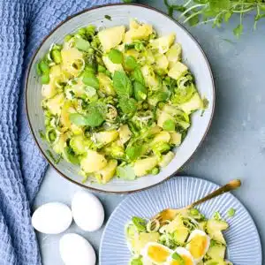 Green potato salad with mint, peas and leek in blue bowl and plate with eggs.