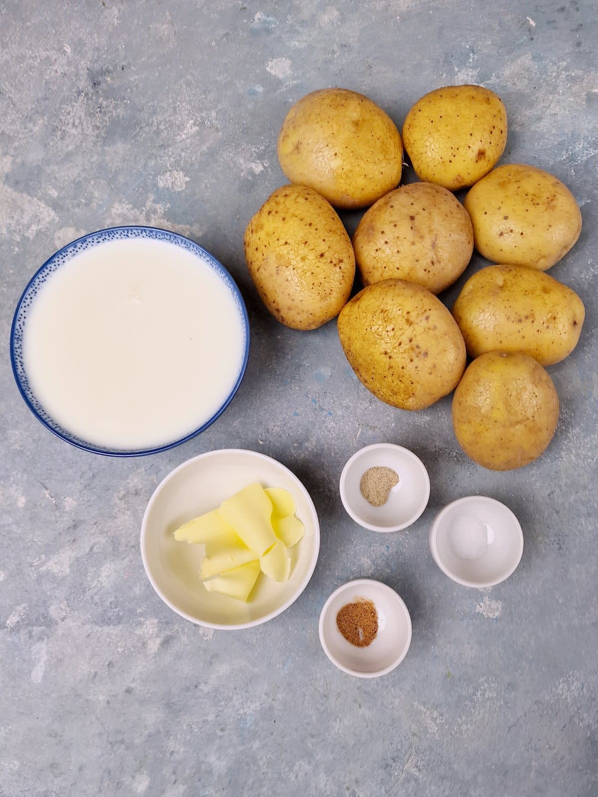 ingredients for mash on blue surface. Large potatoes, milk in bowl, butter and spices. 
