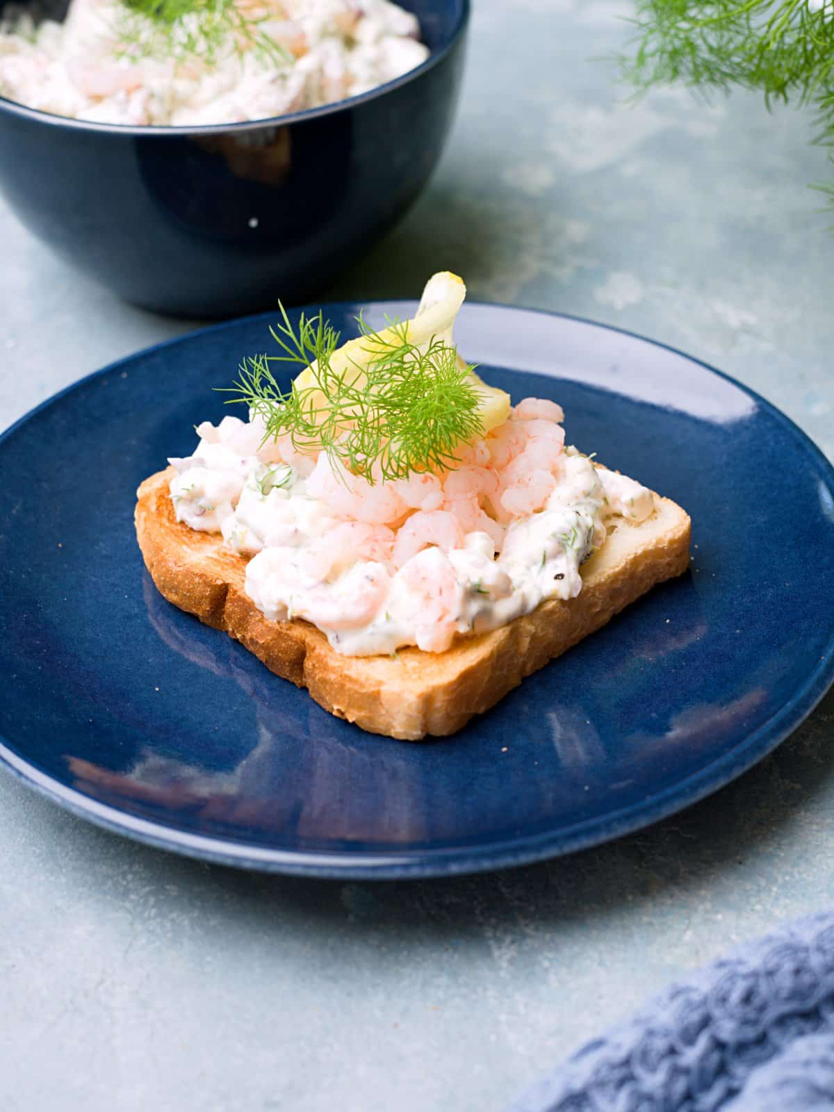 Roasted slice of light toast with some shrimp salad decorated with shrimps, lemon slice and dill on blue plate. 