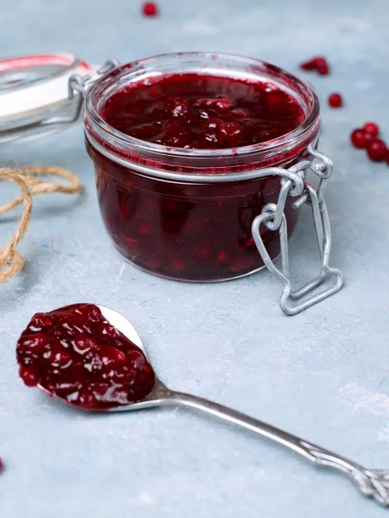 Closeup on a spoon with lingonberry jam and a jar with a plastic ring seal. 