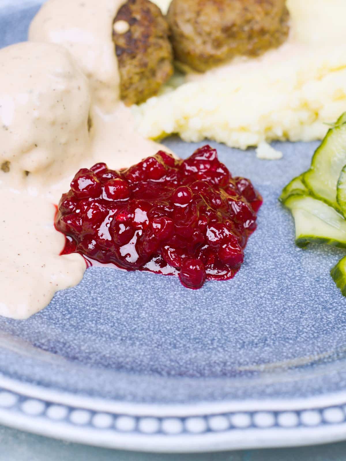 A plate woth meatballs, creamy sauce, mash, cucumbers and lingonberry jam in the forground. 