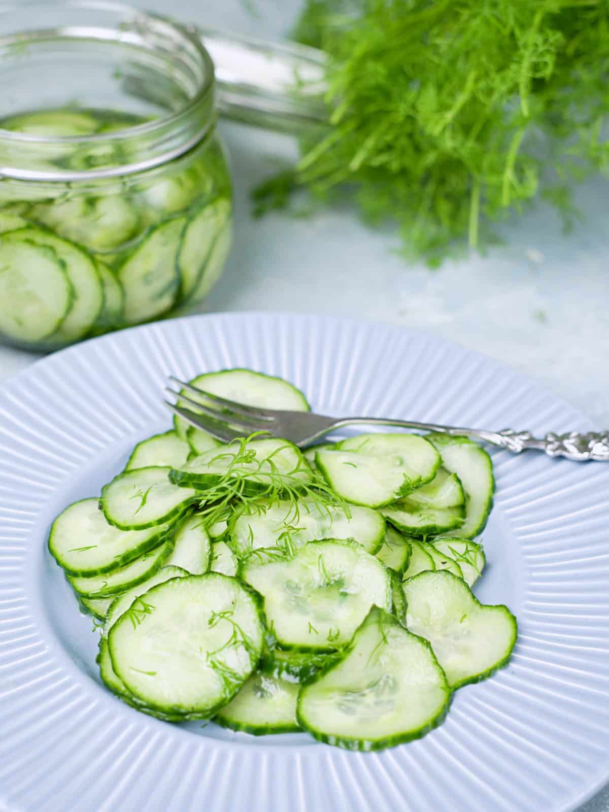 Cucumber slices with dill on a blue plate and a jar with pickled cucumbers in background. 