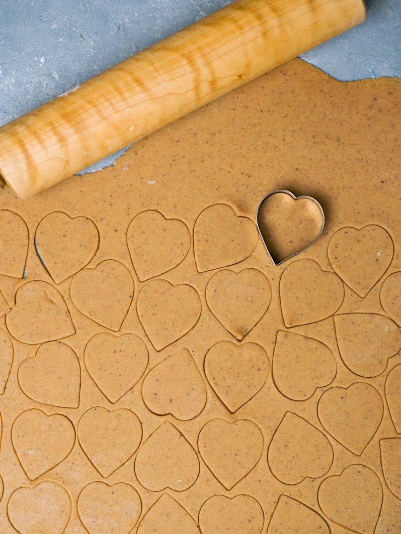Rolled out dough on blue surface with a rolling pin and heart shaped cut forms on it. 