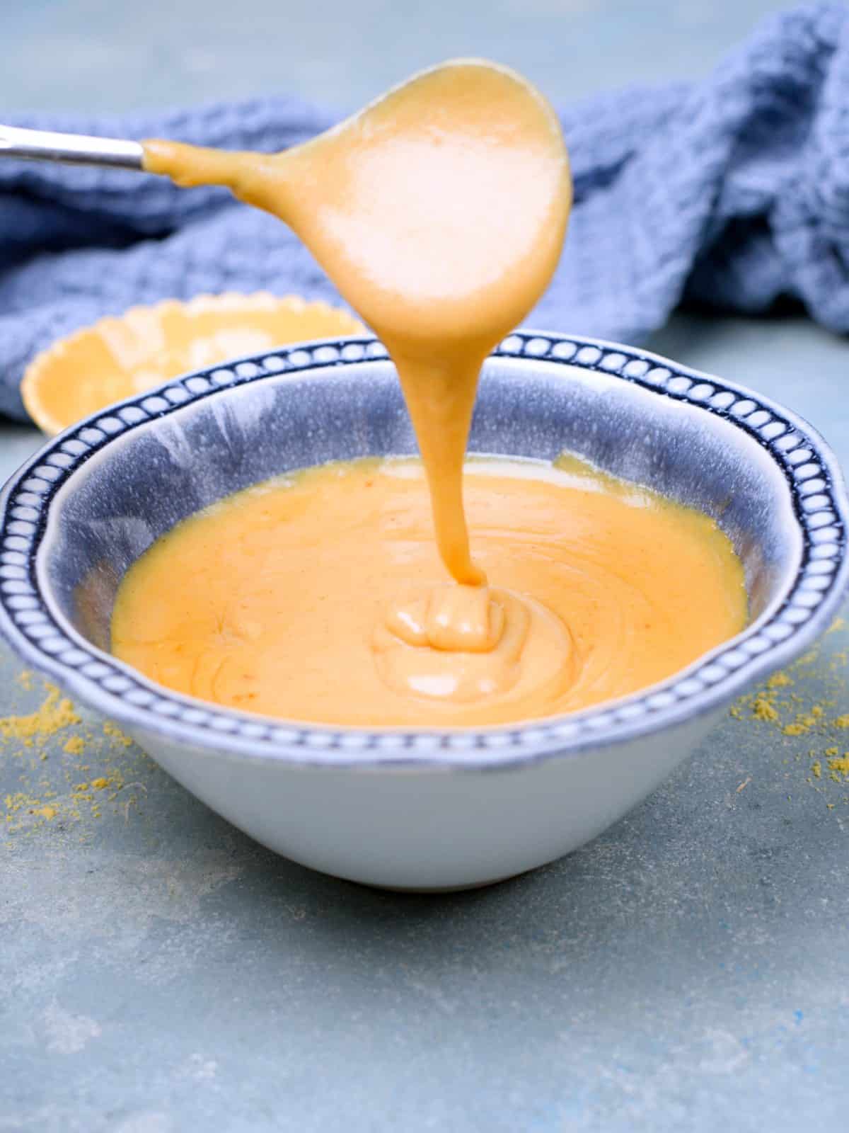 Mustard in a blue romantic bowl and spoon pouring it from the air showing the consistency. Some yellow mustard powder decorates the blue background,.