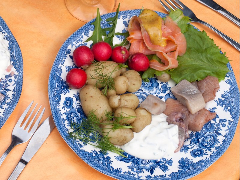 A blu plate served with small new potatoes in dill, herring pieces, yogurt sauce, some low and mustard dressing, radishes and salad. 