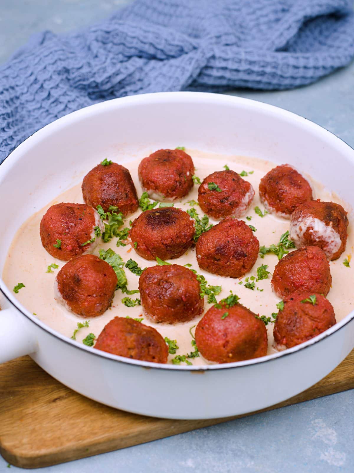 Vegan round fried meatballs lying in cream sauce in a white pan decorated with parsley. 