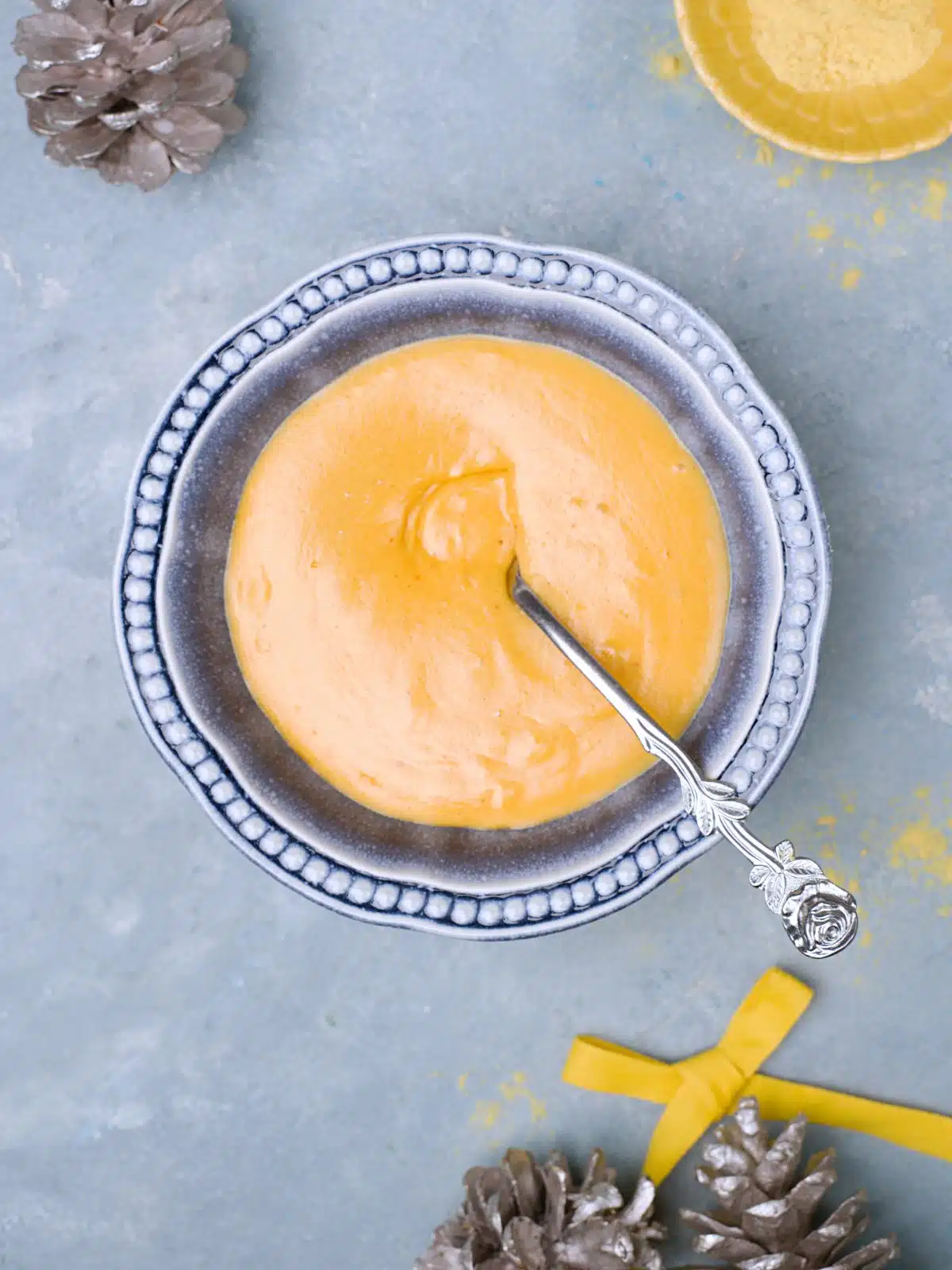 From above picture of  yellow mustard in blue bowl, some silvery pine cones as decor in background. And a yellow ribbon. 